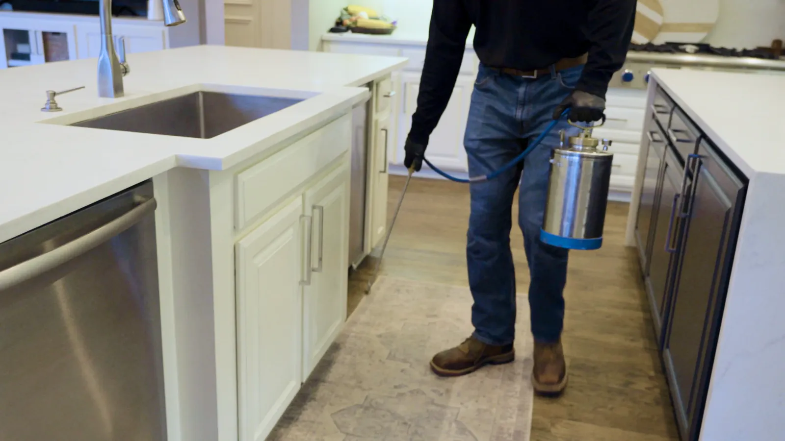 Pest control technician applying treatment along a kitchen baseboard and cabinet edge -- indoor ant bait placement